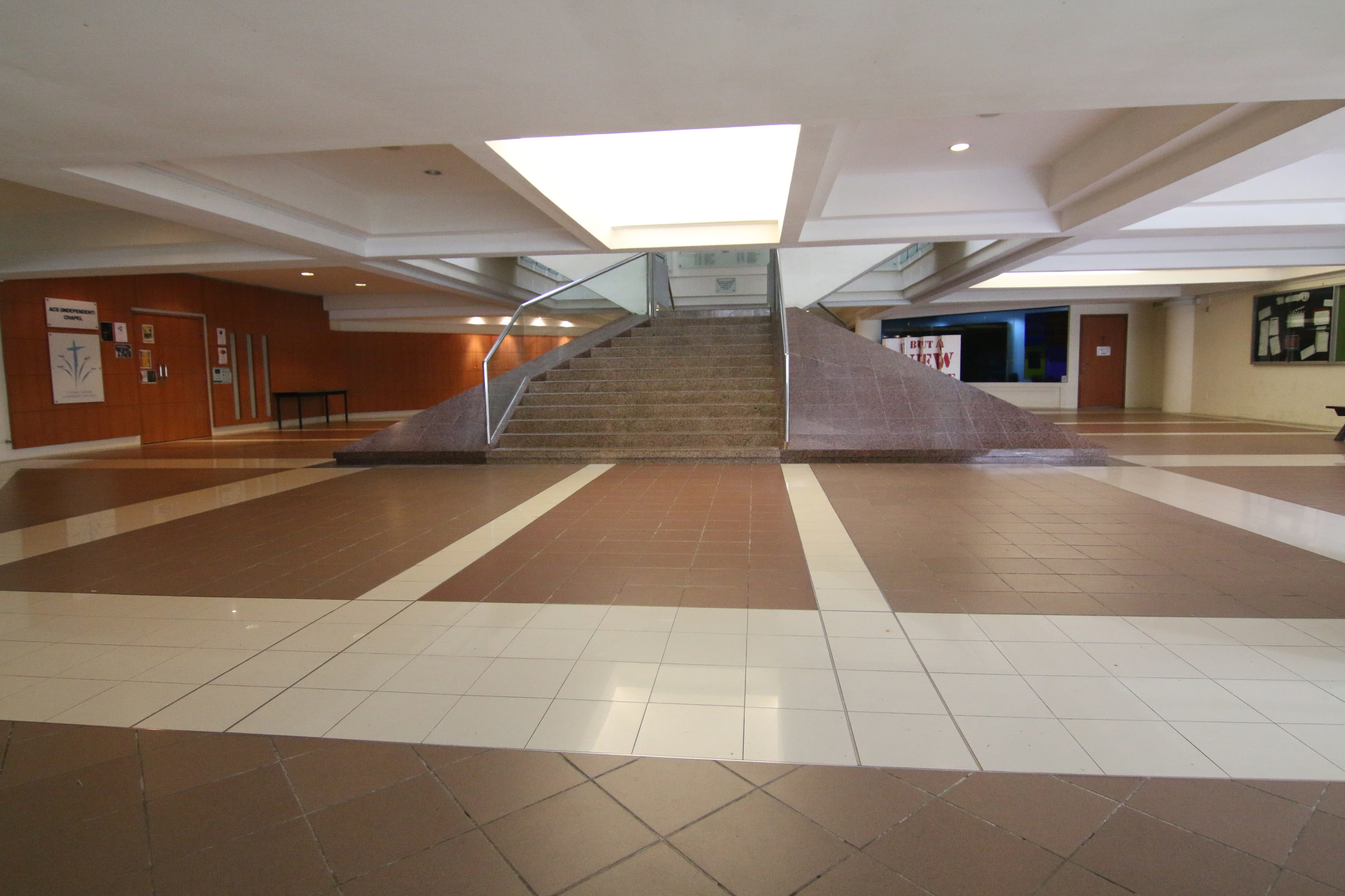 A spacious empty indoor lobby with tiled floors, a central staircase, and skylights above.